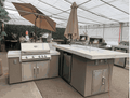 modern outdoor BBQ island with built-in grill, refrigerator, bar counter, fire pit, and umbrellas in a showroom